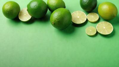The primary subject of the image is a group of limes, which are green and placed on a green surface. The setting is a table with a green background, and the visual style is a photo. The limes are the main focus of the image, and their vibrant green color creates a fresh and lively atmosphere.
