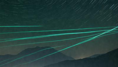 The primary subject of the image is a mountain range with a starry night sky in the background. The setting is a nighttime landscape, and the visual style appears to be a combination of photo and digital art. The colors and mood of the image are predominantly dark and moody, with the green lights adding a touch of whimsy to the scene.