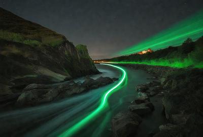 The primary subject of the image is a glowing green light that appears to be coming from a river. The light is shining through the water, creating a mesmerizing effect. The setting is a rocky riverbed, with rocks and a hillside visible in the background. The visual style of the image is a combination of a photo and digital art, as the green light seems to be digitally manipulated to create a unique and captivating scene. The colors and mood of the image are predominantly green, with