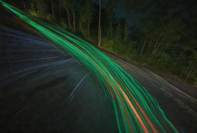 The primary subject of the image is a green light streak, which appears to be a neon light or a light source moving quickly. The setting is a road with trees in the background, creating a nighttime atmosphere. The visual style is a close-up of the light streak, emphasizing its movement and color. The colors and mood of the image are predominantly dark and mysterious, with the green light streak adding a touch of vibrancy and energy to the scene.