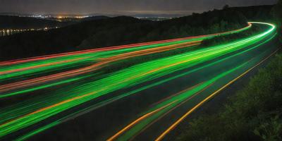 The primary subject of the image is a green light streak, which appears to be a neon light or a green laser beam. The setting is a road or highway, with a dark background. The visual style is a close-up of the green light streak, creating a striking and unique visual effect. The colors and mood of the image are predominantly green, giving it a futuristic and dynamic appearance.