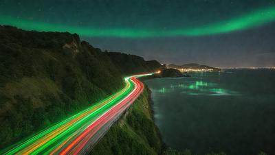 The primary subject of the image is a green light streak, which appears to be a light trail or a green neon light. The setting is a nighttime scene with a bridge and a road, with a mountain in the background. The visual style is a nighttime photo, and the colors and mood are predominantly green, creating a serene and atmospheric scene.