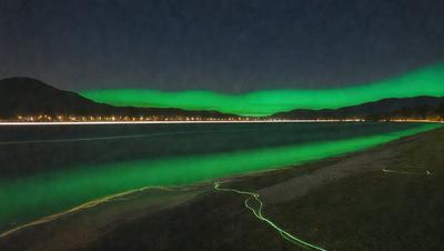 The primary subject of the image is a green glowing light, which appears to be a neon light or a light source with a green hue. The light is located near the water, possibly on a beach or near the ocean. The setting is a nighttime scene, with the light illuminating the area. The visual style of the image is a close-up of the green light, emphasizing its glow and creating a unique and captivating atmosphere. The colors and mood of the image are predominantly green