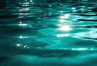 The primary subject of the image is a body of water, which appears to be a large pool of blue water. The water is illuminated by sunlight, creating a bright and serene atmosphere. The setting is outdoors, with the water being the main focus of the scene. The visual style of the image is a photo, capturing the natural beauty of the water and its surroundings. The colors and mood of the image are predominantly blue, with the sunlight adding a touch of warmth and brightness
