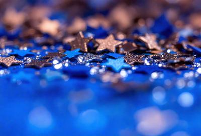 The primary subject of the image is a collection of blue and black star-shaped sequins. The sequins are scattered across a blue background, creating a visually appealing and colorful scene. The visual style of the image is a close-up of the sequins, showcasing their intricate details and patterns. The colors and mood of the image are predominantly blue and black, giving it a vibrant and sophisticated appearance.