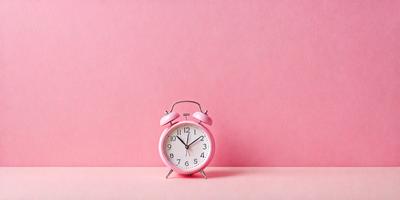 The primary subject of the image is a pink alarm clock. The clock is placed on a pink surface, which could be a table or a wall. The visual style of the image is a photo, capturing the alarm clock in its natural setting. The colors and mood of the image are predominantly pink, creating a warm and inviting atmosphere.