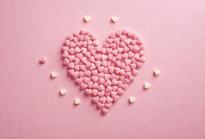 The primary subject of the image is a heart-shaped arrangement of pink and white candy hearts. The heart is made up of numerous small candy hearts, creating a visually appealing and playful display. The setting is a pink background, which complements the colorful candy hearts. The visual style is a combination of digital art and illustration, as the heart is skillfully crafted and arranged to resemble a real heart. The colors and mood of the image are predominantly pink and white,