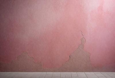 The primary subject of the image is a pink wall with a crack in it. The setting is a room with a white floor. The visual style is a photo, and the dominant colors are pink and white. The overall atmosphere is somewhat distressed or damaged, possibly due to the crack in the wall.