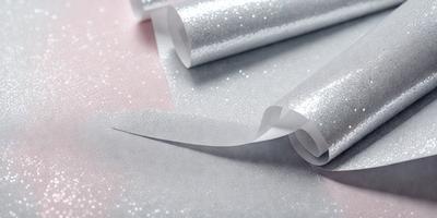 The primary subject of the image is a roll of silver metallic paper, which is shiny and reflective. The paper is unrolled and appears to be a decorative material. The setting is a table, and the visual style is a close-up of the paper. The colors and mood of the image are predominantly silver and reflective, giving it a shiny and metallic appearance.
