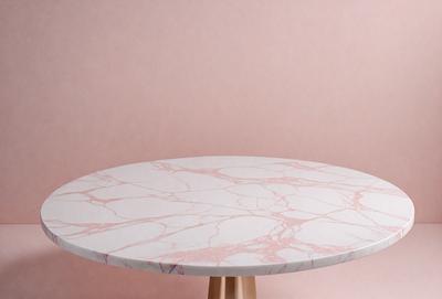 The primary subject of the image is a round table with a marble top. The table is placed on a wooden stand, and it is situated against a pink wall. The setting appears to be a room with a pink background, which adds a warm and inviting atmosphere to the space. The visual style of the image is a close-up of the table, showcasing its marble surface and wooden stand.