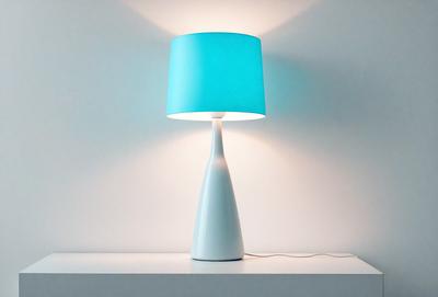 The primary subject of the image is a white lamp with a blue shade. The lamp is placed on a white table, which is situated on a white wall. The visual style of the image is a photo, and the dominant colors are white and blue. The overall atmosphere of the image is clean and minimalistic.