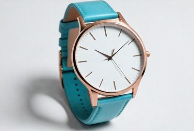 The primary subject of the image is a blue watch with a white face. The watch is placed on a white surface, which serves as the background. The visual style of the image is a photo, capturing the details of the watch and its surroundings. The colors in the image are predominantly blue and white, creating a clean and minimalist atmosphere.
