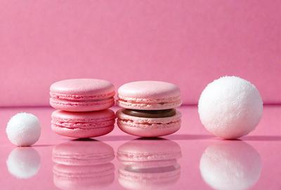 The primary subject of the image is a close-up of two pink macarons with chocolate filling, placed on a pink surface. The setting is a pink background, which adds a vibrant and feminine touch to the scene. The visual style is a photo, capturing the details and textures of the macarons and the pink surface. The colors and mood of the image are predominantly pink, creating a warm and inviting atmosphere.