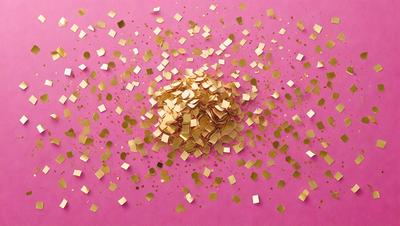 The primary subject of the image is a pile of gold confetti, which is scattered across a pink surface. The setting appears to be a celebration or event, as the confetti is a common decoration for such occasions. The visual style of the image is a close-up of the confetti, showcasing its details and texture. The colors and mood of the image are predominantly pink and gold, creating a warm and festive atmosphere.