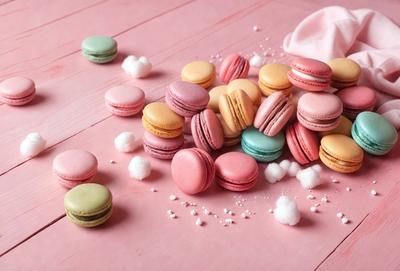 The primary subject of the image is a pink table with a variety of macarons and powdered sugar scattered across it. The table appears to be a dining table, and the macarons are arranged in different shapes and sizes. The setting is a kitchen or dining area, and the visual style is a close-up of the table with the macarons and powdered sugar. The colors and mood of the image are predominantly pink, which gives it a warm and inviting atmosphere.