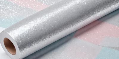 The primary subject of the image is a roll of silver glitter paper. The paper is shiny and metallic, giving it a reflective and glittery appearance. The setting is a table, and the visual style is a close-up of the paper roll. The colors and mood of the image are predominantly silver and reflective, creating a sense of shine and glitter.