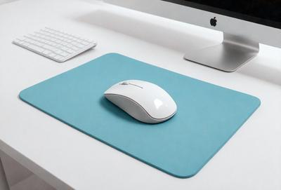 The primary subject of the image is a white computer mouse sitting on a blue mouse pad. The mouse pad is placed on a white desk. The setting is an office environment, with a keyboard and a monitor visible in the background. The visual style is a photo, capturing the details of the mouse and the mouse pad. The colors and mood of the image are predominantly white and blue, creating a clean and organized appearance.