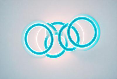 The primary subject of the image is a lighted display featuring three blue rings and one white ring. The rings are arranged in a circular pattern, with the blue rings being positioned around the white ring. The setting appears to be a white wall, which serves as the background for the display. The visual style of the image is a combination of digital art and lighting, creating a unique and eye-catching presentation. The colors and mood of the image are predominantly blue and white, giving it a clean and modern appearance.