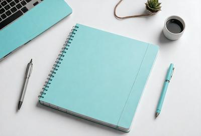 The primary subject of the image is a notebook with a blue cover, placed on a white surface. The notebook is open, revealing a blank page. The setting is a desk with a cup and a potted plant nearby. The visual style is a photo, and the colors and mood are predominantly blue, creating a calm and organized atmosphere.