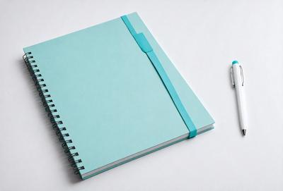 The primary subject of the image is a blue spiral notebook with a pen sitting next to it. The setting is a white table, and the visual style is a photo. The dominant colors are blue and white, creating a calm and organized atmosphere.