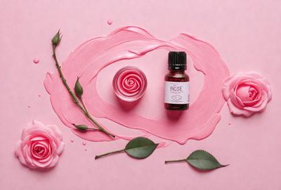 The primary subject of the image is a bottle of rose oil, which is placed on a pink surface. The setting is a pink background, and the visual style is a close-up of the bottle. The dominant colors in the image are pink and red, creating a warm and inviting atmosphere.