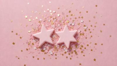 The primary subject of the image is a pair of pink star-shaped candies. They are placed on a pink background, which adds a warm and inviting atmosphere to the scene. The visual style of the image is a close-up, showcasing the details of the candies. The colors and mood of the image are predominantly pink, creating a sense of sweetness and playfulness.