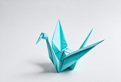 The primary subject of the image is a blue paper crane, which is a traditional Japanese art form. The crane is sitting on a white surface, which serves as the setting or background. The visual style of the image is a close-up of the paper crane, showcasing its intricate details. The colors and mood of the image are predominantly blue and white, giving it a calm and serene atmosphere.