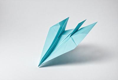 The primary subject of the image is a blue paper airplane. The setting is a white surface, which provides a clean and uncluttered backdrop for the airplane. The visual style is a close-up of the paper airplane, showcasing its details and texture. The colors and mood of the image are predominantly blue, giving it a calm and serene atmosphere.