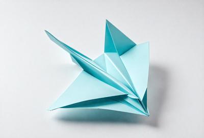 The primary subject of the image is a blue paper kite. The kite is sitting on a white surface, which serves as the background. The visual style of the image is a photo, capturing the details of the kite. The colors and mood of the image are predominantly blue, giving it a calm and serene atmosphere.