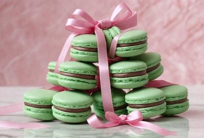 The primary subject of the image is a stack of green macaroon cookies. The cookies are arranged in a pile, with some of them being decorated with pink ribbons. The setting is a table, and the visual style is a close-up of the cookies. The colors and mood of the image are predominantly green and pink, creating a visually appealing and inviting scene.