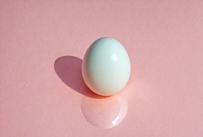 The primary subject of the image is a white egg, which is placed on a pink surface. The egg is the focal point of the scene, and it appears to be sitting on a piece of glass or a reflective surface. The setting is a simple, clean environment, with no other objects or distractions visible in the image. The visual style is a close-up of the egg, emphasizing its shape and texture. The colors in the image are predominantly pink and white, creating a calm and serene atmosphere