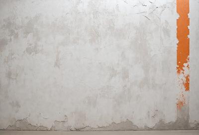 The primary subject of the image is a white wall with a peeling paint surface. The setting is a room with a bare, unadorned appearance. The visual style is a close-up of the wall, showcasing the peeling paint and the texture of the wall. The colors and mood of the image are predominantly white, with a slightly aged and worn appearance.