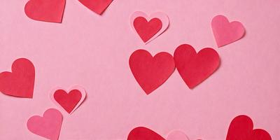 The primary subject of the image is a collection of red hearts, which are arranged on a pink background. The hearts are made of paper and are placed in various positions, creating a visually appealing and playful scene. The setting is a pink surface, which adds to the overall mood and color scheme of the image. The visual style of the image is a combination of digital art and illustration, as the hearts are created using paper and then digitally arranged on the pink background.