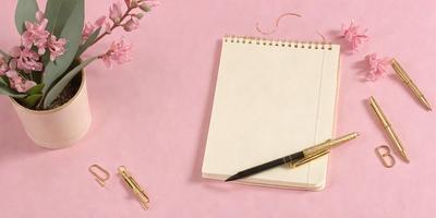 The primary subject of the image is a notebook with a pen on top of it. The setting is a pink table, and the visual style is a photo. The colors and mood of the image are predominantly pink, creating a warm and inviting atmosphere.