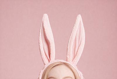 The primary subject of the image is a woman wearing a pink bunny costume. The setting is a pink background, which creates a warm and playful atmosphere. The visual style is a close-up of the woman's face, emphasizing her expression and the costume. The colors and mood are predominantly pink, which adds a sense of fun and lightheartedness to the scene.