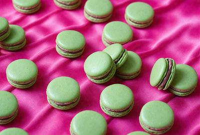 The primary subject of the image is a close-up of a green macaron, which is placed on a pink surface. The setting is a table with a pink cloth, and the visual style is a photo. The dominant colors are green and pink, creating a vibrant and appetizing atmosphere.