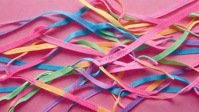 The primary subject of the image is a close-up of a colorful ribbon. The ribbon is made up of multiple colors, including pink, purple, blue, green, yellow, and orange. The ribbon is woven and appears to be a decorative item. The setting is a pink surface, which provides a contrasting background for the vibrant ribbon. The visual style of the image is a close-up, focusing on the details of the ribbon. The colors and mood of the