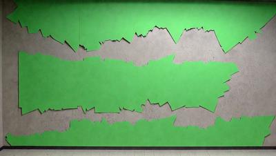 The primary subject of the image is a green wall with a large, intricate design on it. The design appears to be a combination of a mountain and a wave, giving the impression of a mountain wave. The setting is indoors, with the wall being the main focus of the scene. The visual style of the image is a mix of digital art and illustration, creating a unique and eye-catching design. The dominant colors in the image are green and grey, giving the wall a calm and serene atmosphere.
