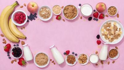 The primary subject of the image is a pink table with various food items and drinks arranged on it. The table is set with a bowl of cereal, a bowl of granola, a bowl of fruit, and a bowl of yogurt. There are also two bottles of milk on the table. The setting is a dining table, and the visual style is a realistic representation of the food items and table. The colors and mood of the image are warm and inviting, with the p