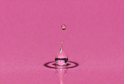 The primary subject of the image is a single drop of water, which is captured in mid-air. The setting is a pink background, which adds a vibrant and warm atmosphere to the scene. The visual style is a close-up of the water drop, emphasizing its details and texture. The colors and mood of the image are predominantly pink, creating a sense of warmth and softness.