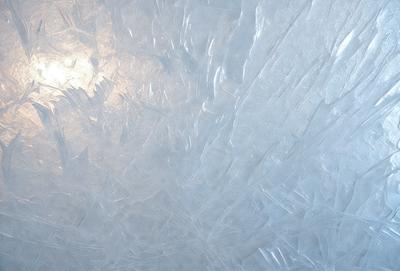 The primary subject of the image is a large, frosty, and icy surface. The setting is a snowy environment, with a bright sun shining on the frozen surface. The visual style is a combination of photo and digital art, creating a unique and captivating scene. The dominant colors are white and blue, with the frosty surface being predominantly white and the sunlight creating a blue hue. The overall atmosphere is cold and serene, with the sunlight creating a sense of warmth and life amidst