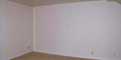 The primary subject of the image is a white wall with a power outlet on it. The setting is a room with a carpeted floor. The visual style is a photo, and the colors and mood are predominantly white, giving the room a clean and minimalist appearance.