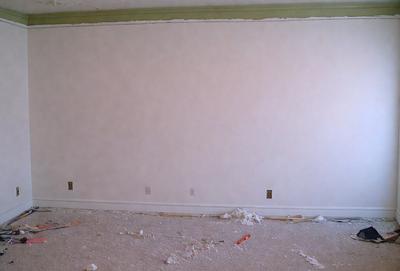 The primary subject of the image is a large, empty white wall. The setting is a room with a wooden floor, and the visual style is a photo. The colors and mood of the image are predominantly white, giving it a clean and minimalist appearance.