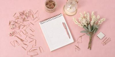 The primary subject of the image is a notebook with a pink cover, placed on a pink surface. The setting is a table with a vase and a cup nearby. The visual style is a photo, and the colors and mood are predominantly pink, creating a warm and inviting atmosphere.