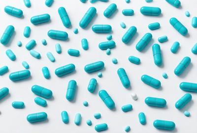 The primary subject of the image is a large number of blue pills arranged in a visually appealing manner. The setting is a white background, which provides a clean and uncluttered look. The visual style is a combination of a photo and digital art, creating a unique and eye-catching display. The dominant colors are blue and white, giving the image a calm and soothing atmosphere.