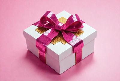 The primary subject of the image is a pink bow-shaped ribbon tied around a white box. The setting is a pink background, which adds a warm and inviting atmosphere to the scene. The visual style is a close-up of the box and bow, emphasizing the details of the ribbon and the box. The colors and mood of the image are predominantly pink, creating a sense of love and affection.