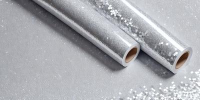 The primary subject of the image is a roll of silver glitter paper. The paper is shiny and reflective, giving it a metallic appearance. The setting is a table, and the visual style is a close-up of the paper. The colors and mood of the image are predominantly silver and reflective, creating a shiny and glittery atmosphere.