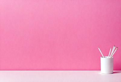 The primary subject of the image is a white pencil placed on a white surface, which appears to be a table. The setting is a pink wall, creating a contrasting background. The visual style is a simple, minimalist composition, focusing on the pencil and the wall. The colors and mood of the image are predominantly white and pink, giving it a clean and fresh appearance.