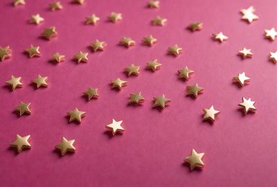 The primary subject of the image is a collection of gold stars scattered across a pink surface. The setting appears to be a pink background, which adds a warm and inviting atmosphere to the scene. The visual style of the image is a close-up of the stars, showcasing their intricate details. The colors and mood of the image are predominantly pink and warm, creating a sense of comfort and relaxation.