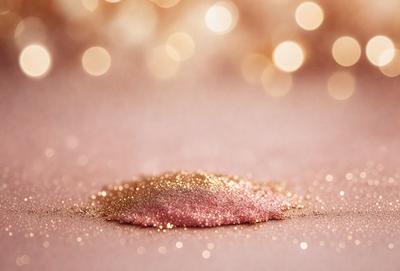 The primary subject of the image is a small pink and gold glittery ball, which is the main focus of the scene. The setting is a pink background, which adds to the overall mood of the image. The visual style is a close-up of the glittery ball, showcasing its details and texture. The colors and mood of the image are predominantly pink and gold, creating a warm and festive atmosphere.